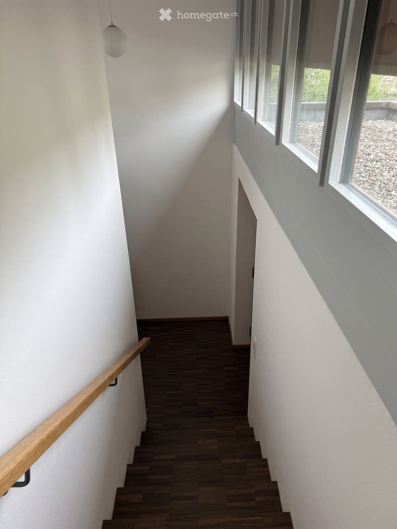 Modern hallway with wooden stairs, white walls, and large windows showing a view of a garden area with gravel.