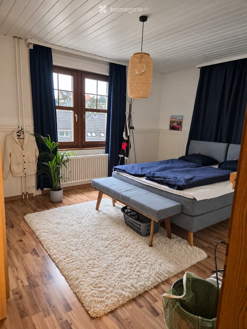 Cozy bedroom with wooden flooring, a large bed with blue bedding, a bench at the foot of the bed, a white shag rug, a green chair, a potted plant, and a coat rack with a jacket. The room features large windows with dark curtains and a hanging woven lamp.