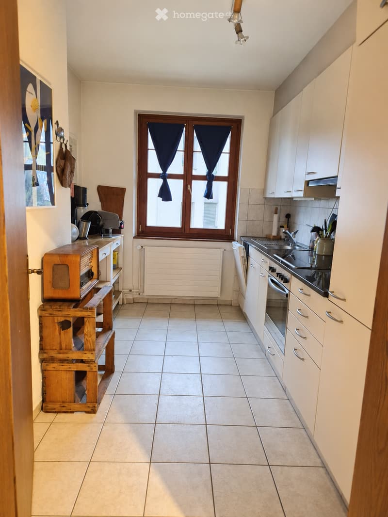 The kitchen features white cabinets, a tiled floor, a window with dark curtains, and a wooden table with a vintage radio on it. There is a sink with a faucet, an oven, and a radiator under the window.