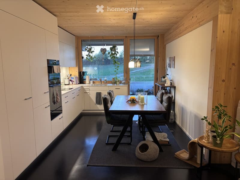 Modern kitchen and dining area with white cabinets, dark flooring, and large windows. Includes a dining table with chairs, a small side table with a potted plant, and a cat bed.
