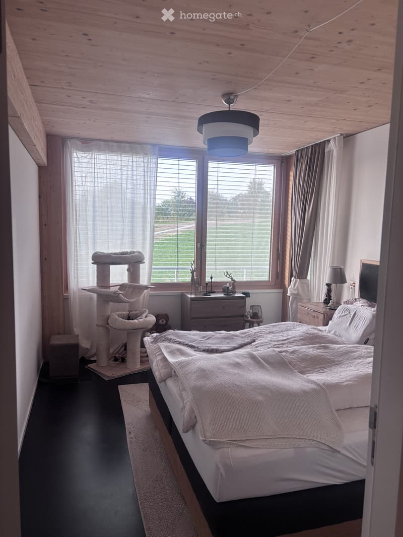 Cozy bedroom with a large bed, cat tree, nightstand, and a view of the outdoors through large windows.