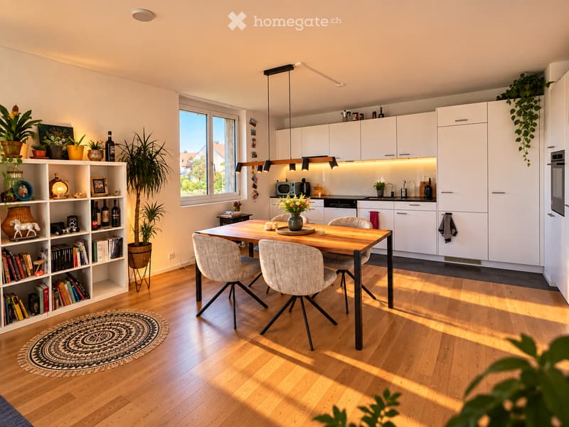 Bright, modern dining and kitchen area with wooden flooring, white cabinets, a wooden dining table with four chairs, a bookshelf with books and decorative items, and a potted plant near the window.