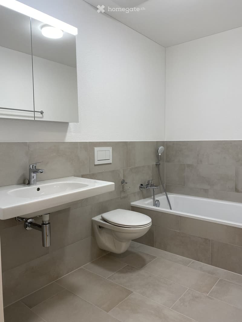 Modern bathroom with a white sink, chrome faucet, toilet, bathtub, and showerhead. Grey tiled walls and beige tiled floor.