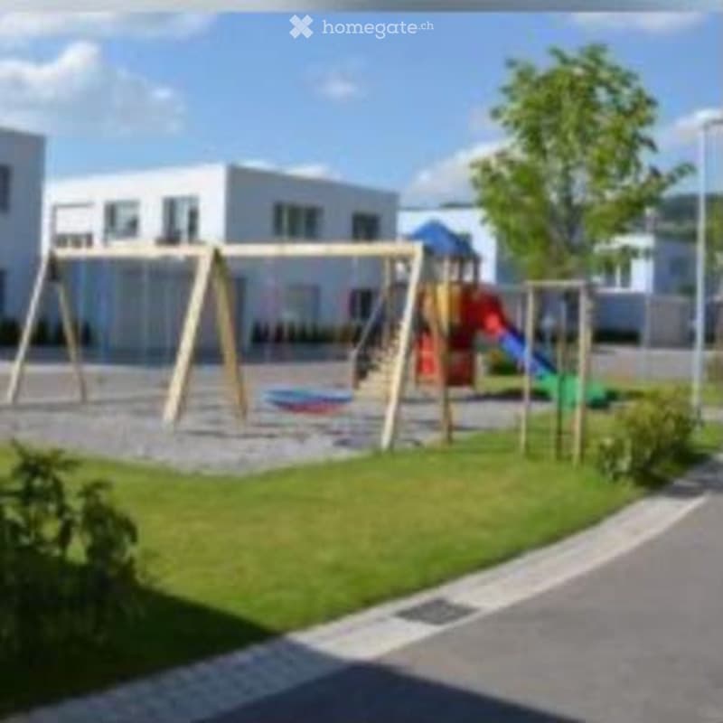 Playground area with swings, slide, and climbing frame, surrounded by grass and a paved path.