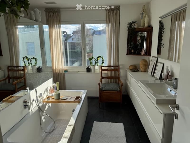Bathroom with a bathtub, sink, two wooden chairs, a mirror, framed picture, vase, and plants on the windowsill.