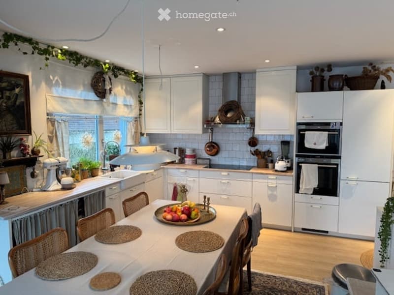 Bright, modern kitchen with white cabinets, a central dining table with chairs, and various appliances including a fridge, oven, and blender. The space features a fruit bowl on the table, potted plants, and a picture frame on the wall.