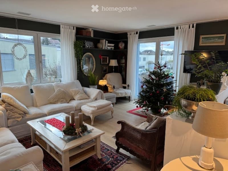 Bright living room with large windows, white sofa, coffee table, ottoman, armchair, floor lamp, table lamp, mirror, television, potted plants, and a Christmas tree.
