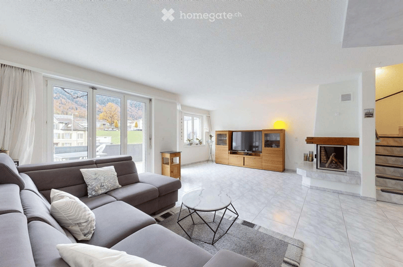Modern living room with large windows offering a scenic view, a grey sectional sofa, a round coffee table, a fireplace, a TV cabinet, and side tables.