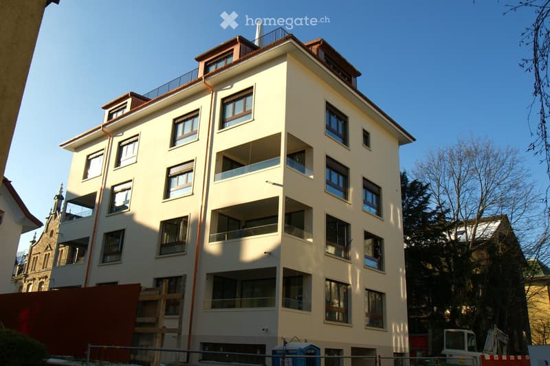 Modern multi-story residential building with light-colored facade, numerous windows, and balconies. The building features a flat roof with a small chimney and is surrounded by trees and other structures.