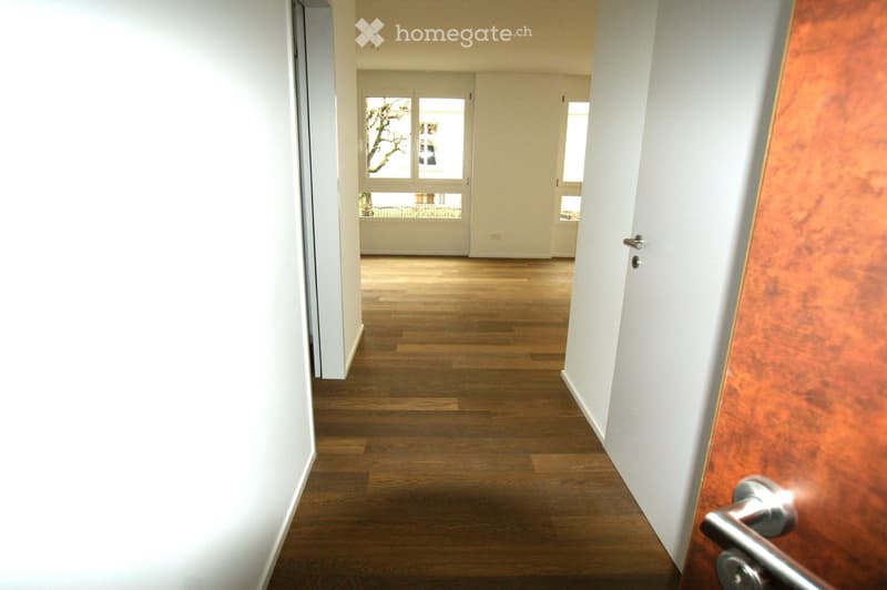 Empty hallway with wooden flooring, white walls, and a door leading to another room.