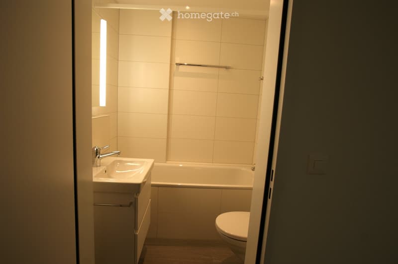 Modern bathroom with white tiles, a sink with a chrome faucet, a bathtub, a toilet, and a built-in cabinet.
