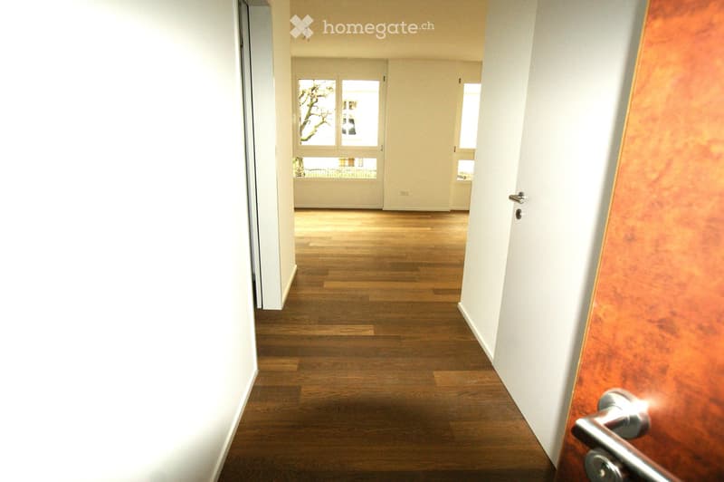 Empty hallway with wooden flooring, white walls, and a door leading to another room.