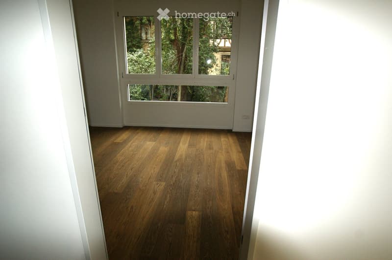 Empty room with wooden flooring and large windows showing greenery outside. No furniture or equipment present.