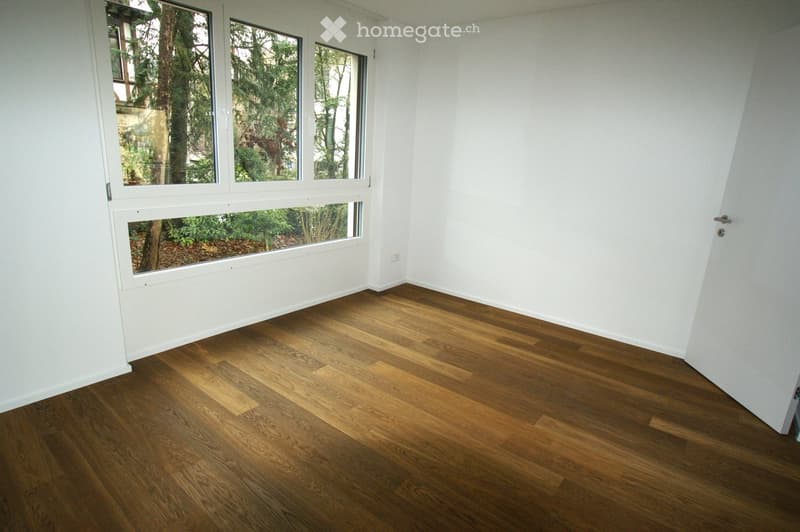 Empty room with light wooden flooring and large windows showing greenery outside. Bare walls and no furniture.