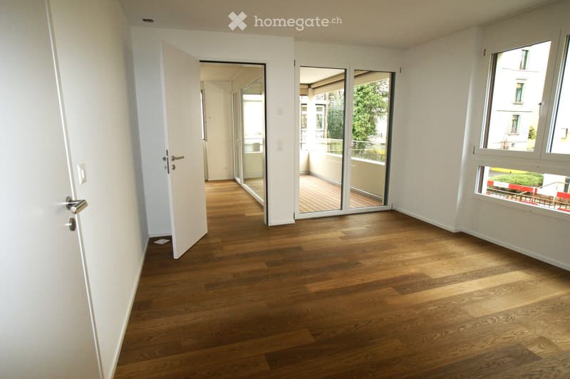 Empty room with wooden flooring, white walls, and large windows. A sliding glass door leads to a balcony area with wooden decking.