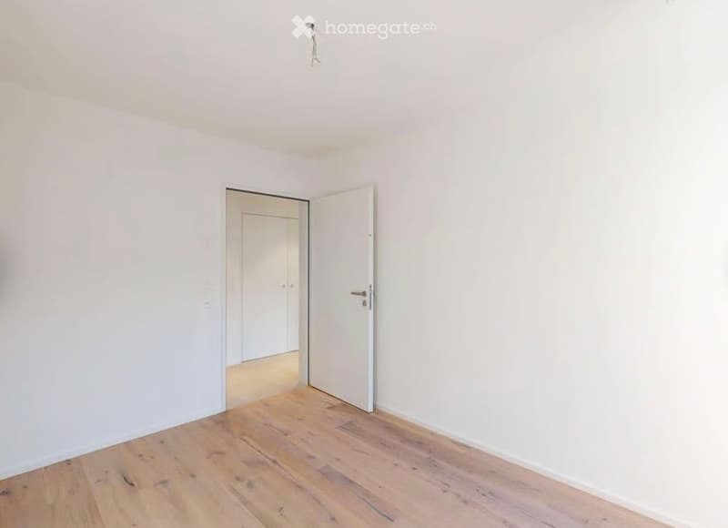 Empty room with light wooden flooring and white walls, featuring an open door leading to another room.