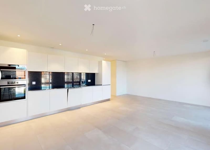 Empty room with white walls and light-colored flooring, featuring a modern kitchen area with white cabinets and stainless steel appliances.