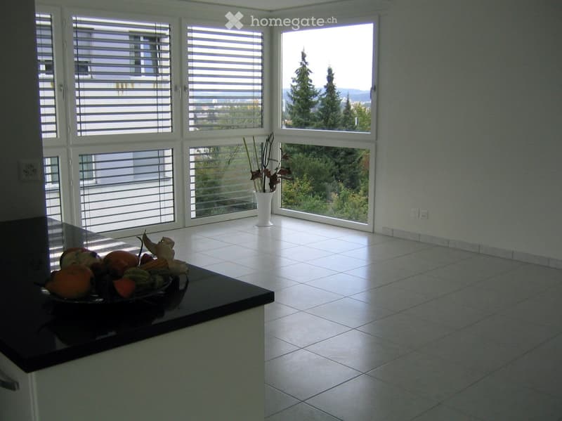 Bright, minimalist room with large windows, white tiled floor, and a small black countertop holding a bowl of fruit. A white vase with dried branches stands near the windows.