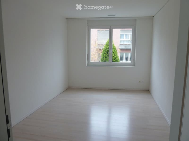 Empty room with light-colored wooden flooring and white walls, featuring a large window with a view of greenery and neighboring buildings.