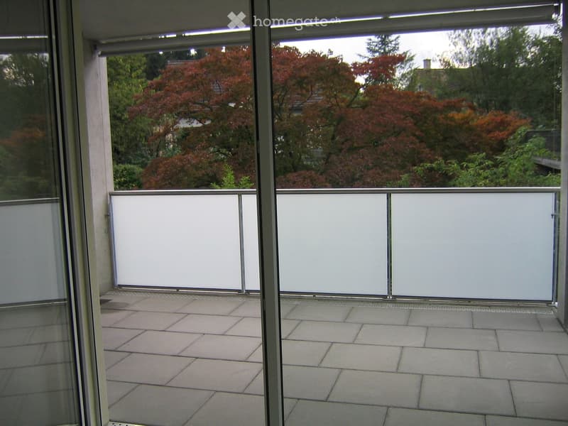 Balcony with tiled floor, white sliding glass doors, and a view of trees and a house in the background.