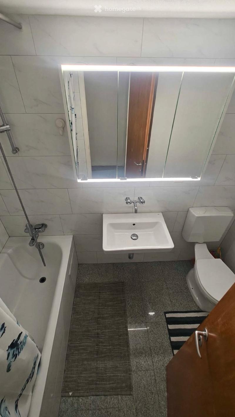 Modern bathroom with a bathtub, sink, toilet, and a large mirror with built-in lighting. The floor is tiled, and the walls are covered with light-colored tiles.