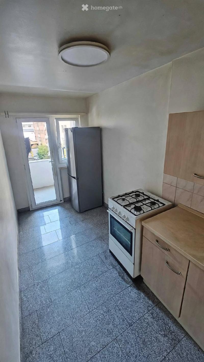 Empty kitchen with tiled floor, white stove, refrigerator, and wooden cabinets. A sliding glass door leads to a balcony.