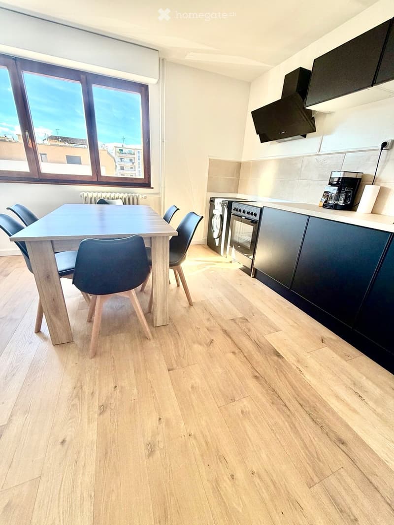 A modern kitchen and dining area with light wooden flooring, a large window offering a city view, a dining table with four chairs, and modern kitchen appliances including an oven, washing machine, and coffee machine.