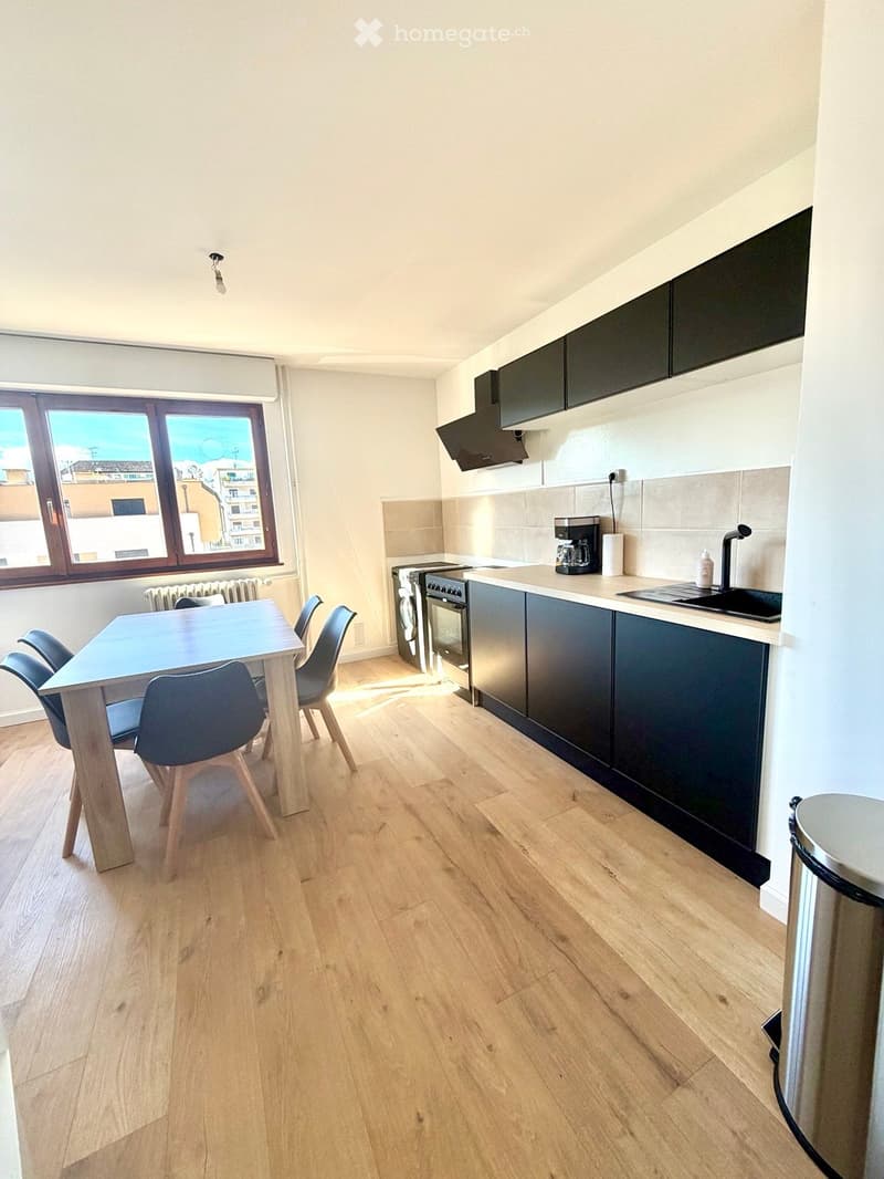 Modern kitchen with wooden flooring, dark cabinets, a dining table with chairs, and appliances including an oven, fridge, and coffee maker.