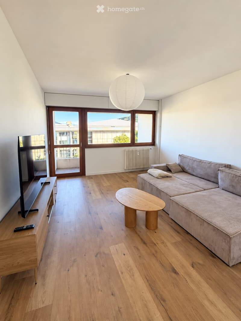 Modern living room with wooden flooring, large windows offering a view, a grey sofa, a round wooden coffee table, a flat-screen TV on a wooden stand, and a remote control on the stand.