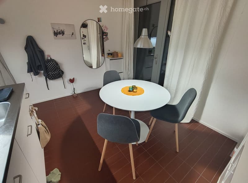A small, modern dining area with a round white table, four grey chairs, a mirror, and a backpack hanging on the wall. The room has a tiled floor and a sink visible on the left side.