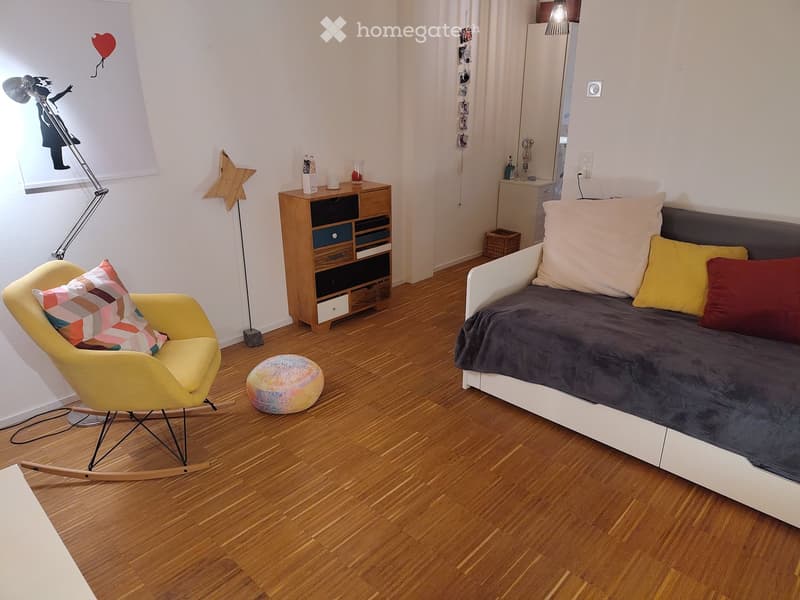 Cozy bedroom with a modern sofa, rocking chair, colorful stool, wooden cabinet, and a floor lamp. The room features wooden flooring and a playful mural on the wall.