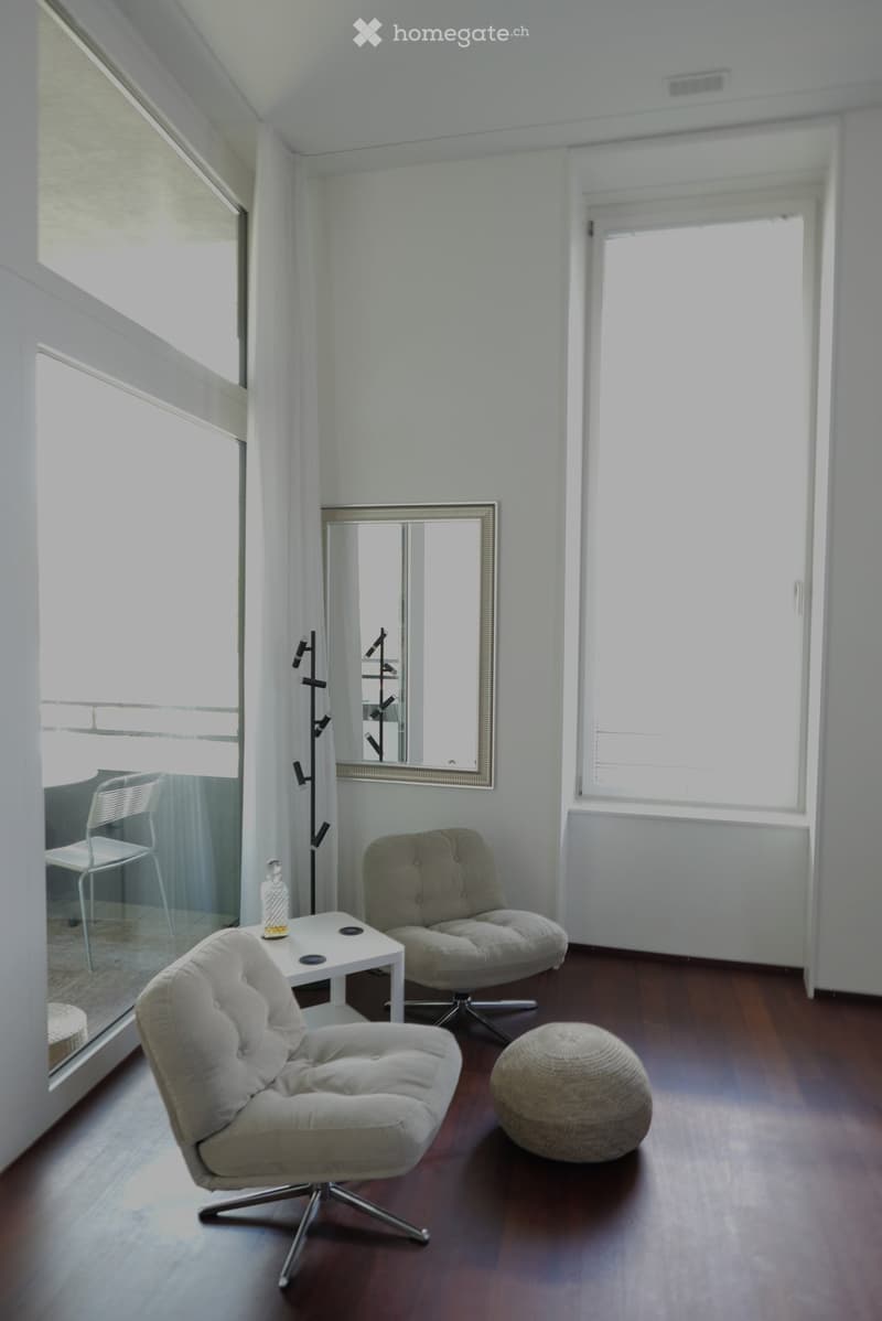 Bright, modern living space with wooden flooring, large windows, and a view. Features include a white table with a decorative bottle, two armchairs, a round ottoman, and a large mirror on the wall.
