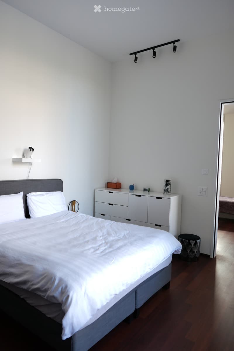 A modern bedroom with a neatly made bed, a white nightstand with a lamp, a small stool, and a mirror on the wall.