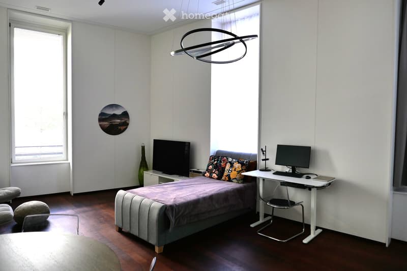 Modern bedroom with a grey upholstered bed, a white desk with a computer, a round mirror, and a decorative vase. The room features large windows and a contemporary pendant light.