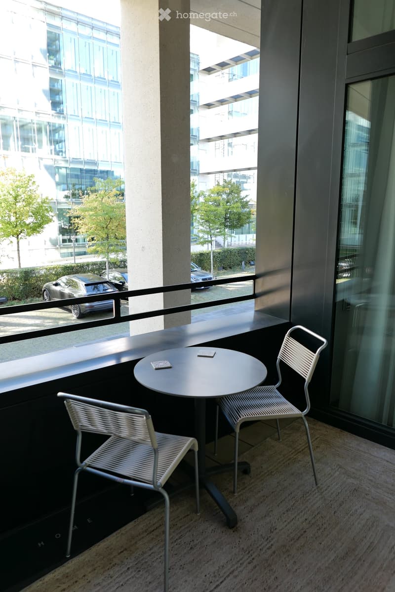 Balcony with a round table and two white chairs, overlooking a modern building and parking area.
