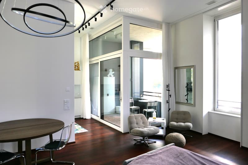 Modern, open-concept living space with a wooden floor, large windows, and a balcony. Features a round wooden table, chairs, a sofa, a mirror, and a stool.