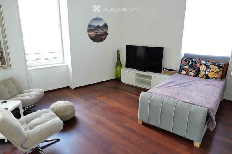 Modern bedroom with a wooden floor, a grey upholstered bed, colorful pillows, a white TV stand, a green vase, a round mirror, and two comfortable chairs.