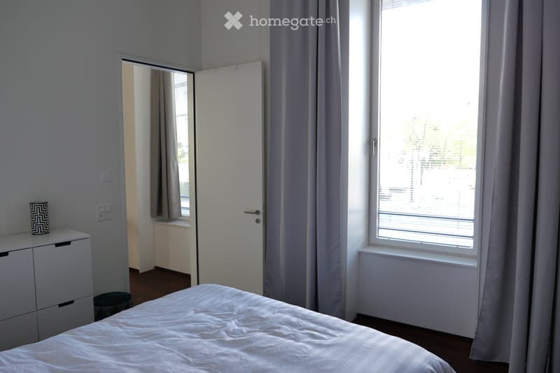 A modern bedroom with a neatly made bed, a white cabinet with a patterned vase, a mirror, and a window with blinds and curtains.
