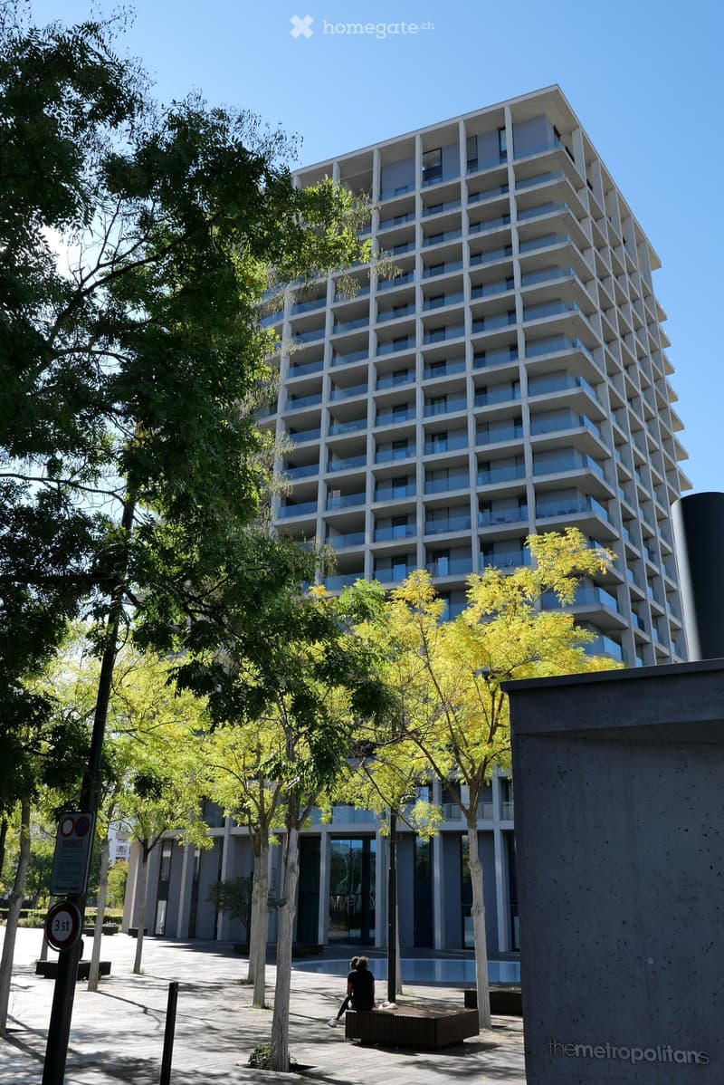 Modern high-rise building with multiple floors, balconies, and large windows, surrounded by trees and a paved area with a bench.