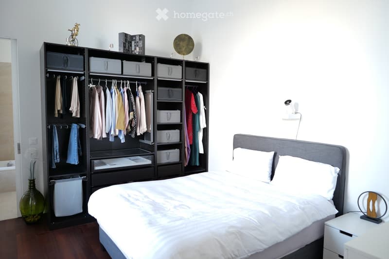Modern bedroom with a large bed, white bedding, a tall black wardrobe with multiple sections, a green vase with dried flowers, and a small nightstand with a lamp.