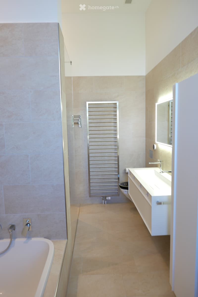Modern bathroom with beige tiles, a bathtub, a sink with a mirror, a radiator, and a toilet.