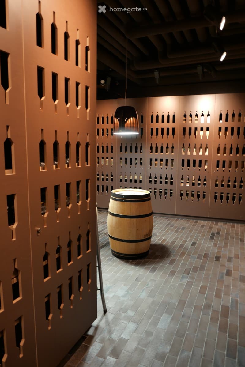 A stylish wine cellar with a large wooden barrel in the center, surrounded by walls lined with wine bottles, illuminated by warm lighting and a modern pendant lamp.