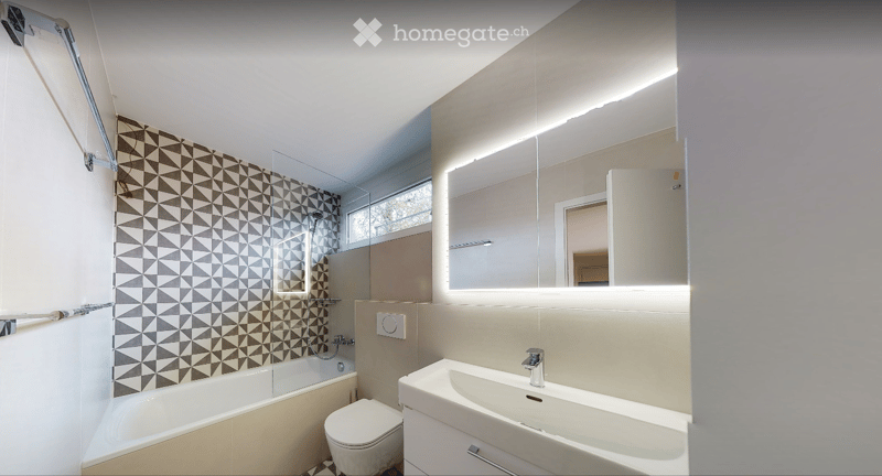 Modern bathroom with a bathtub, toilet, sink, and large mirror. Features geometric patterned tiles on the shower wall and a sleek, contemporary design.