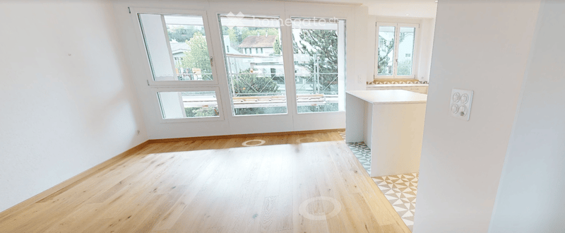 Empty room with wooden flooring, white walls, and large windows offering a view of the outside. A small kitchen counter is visible on the right side.