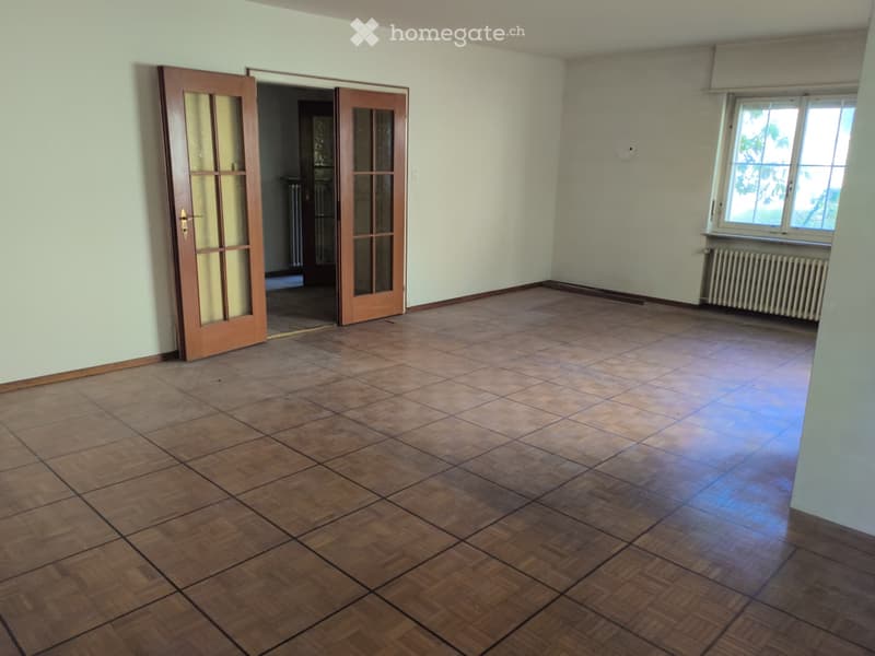Empty room with wooden flooring, light green walls, and large windows with white frames. There is a radiator under the windows and a wooden door with glass panels leading to another room.