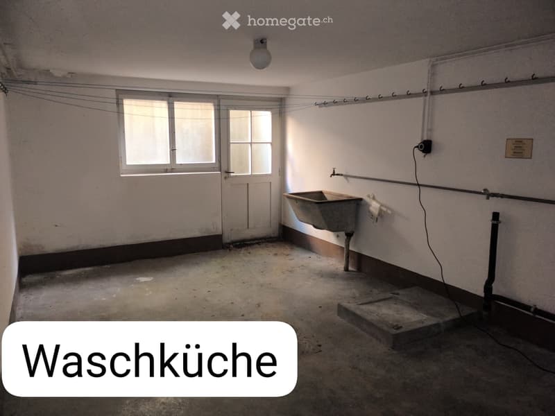 Empty washroom with bare walls, concrete floor, a single window, a door, a sink, and a small utility area with pipes and a plug.