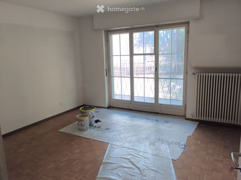 Empty room with wooden flooring, white walls, large sliding glass doors leading to an outdoor area, and a radiator on one wall. The floor is covered with protective sheets and there are a few paint cans and tools visible.