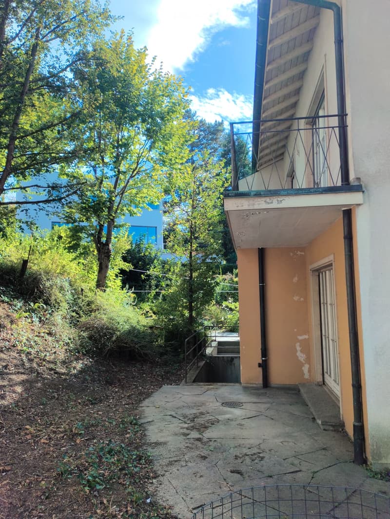 A raised ground floor entrance to a building with a concrete pathway, metal railing, and a small staircase leading up to a balcony area.