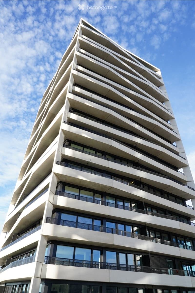 Modern multi-story building with a sleek, contemporary design featuring large windows and balconies, set against a clear blue sky.