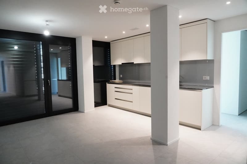 Modern kitchen with white cabinets, a central island, and a large window with blinds. The space features a clean, minimalist design with a tiled floor and recessed lighting.